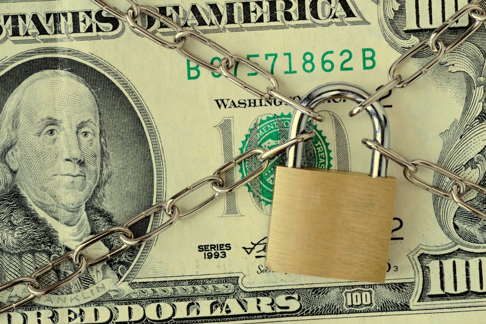 Ein $100 Schein wird durch vier Ketten abgesichert, die in der Mitte in einem Schloss zusammenlaufen.